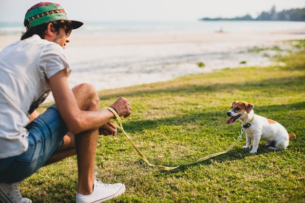 Uomo giovane alla moda hipster camminare e giocare con il cane in spiaggia tropicale