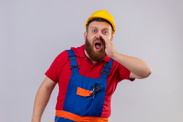 Uomo giovane barbuto costruttore in uniforme da costruzione e casco di sicurezza che grida o chiama qualcuno con la mano vicino alla bocca