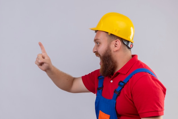 Uomo giovane barbuto costruttore in uniforme da costruzione e casco di sicurezza gridando emotivo e preoccupato che mostra il dito indice