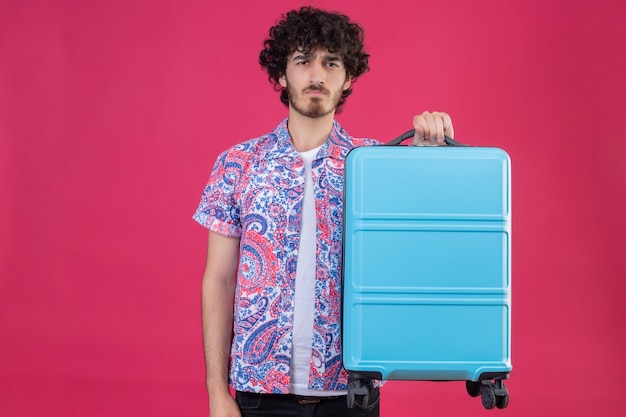 Uomo giovane bello riccio sgradevole del viaggiatore che tiene la valigia sulla parete rosa isolata con lo spazio della copia