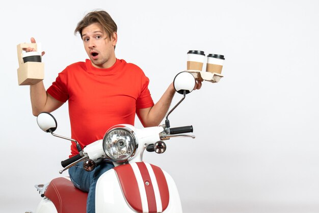 Uomo giovane corriere confuso in uniforme rossa che si siede sullo scooter che tiene gli ordini sulla parete gialla