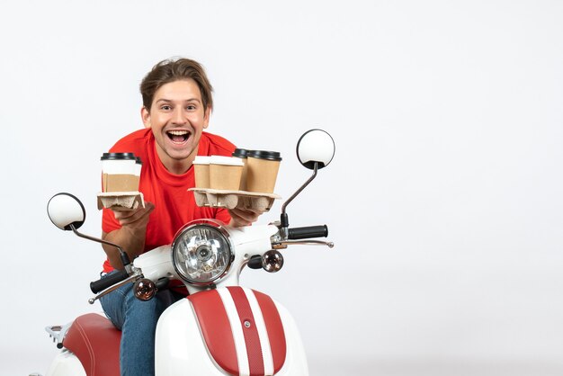 Uomo giovane corriere felice in uniforme rossa che si siede sul motorino che tiene gli ordini sulla parete gialla