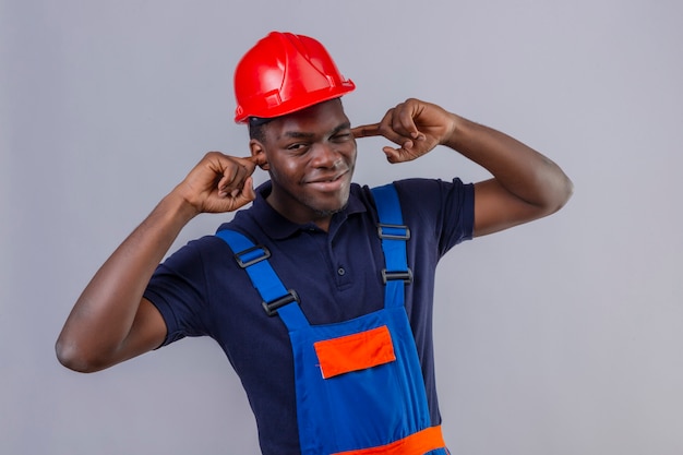 Uomo giovane costruttore afroamericano che indossa uniformi da costruzione e casco di sicurezza coning orecchie con le dita con espressione infastidita per il rumore di suoni forti in piedi