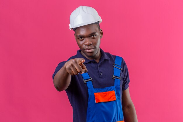 Uomo giovane costruttore afroamericano dispiaciuto che indossa l'uniforme da costruzione e casco di sicurezza che punta il dito con espressione seria sul viso in piedi sul rosa
