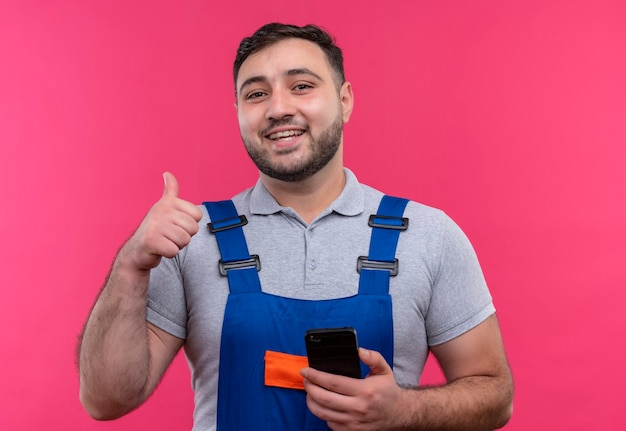 Uomo giovane costruttore in costruzione uniforme tenendo lo smartphone che mostra i pollici in su sorridente