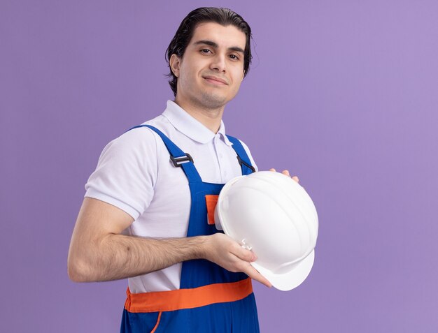 Uomo giovane costruttore in uniforme da costruzione che tiene il suo casco di sicurezza guardando la parte anteriore con espressione fiduciosa in piedi sopra la parete viola