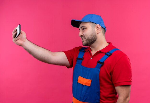 Uomo giovane costruttore in uniforme da costruzione e cappuccio rendendo selfie utilizzando il suo telefono cellulare sorridente