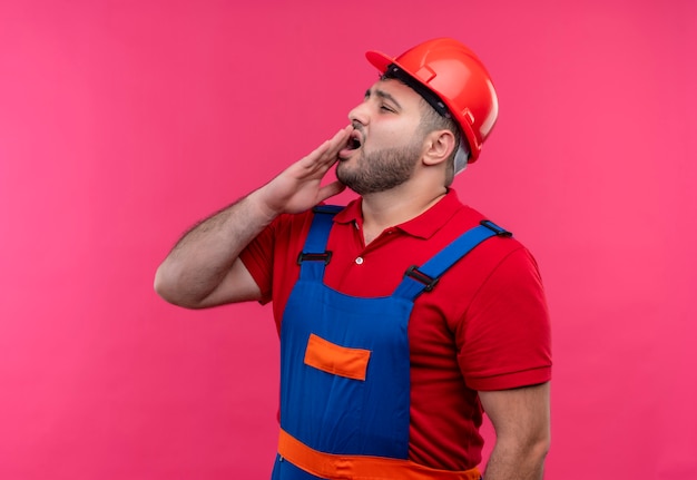 Uomo giovane costruttore in uniforme da costruzione e casco di sicurezza che guarda da parte annoiato chiamando qualcuno con la mano vicino alla bocca