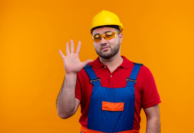 Uomo giovane costruttore in uniforme da costruzione e casco di sicurezza che guarda l'obbiettivo che fluttua con la mano sorridente