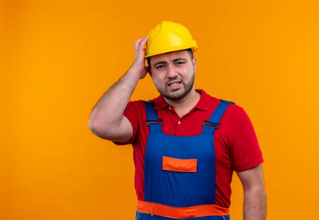 Uomo giovane costruttore in uniforme da costruzione e casco di sicurezza che guarda l'obbiettivo scontento con la mano sulla testa per errore che sembra confuso