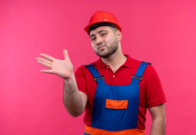 Uomo giovane costruttore in uniforme da costruzione e casco di sicurezza che guarda l'obbiettivo sorridente gesticolando con la mano