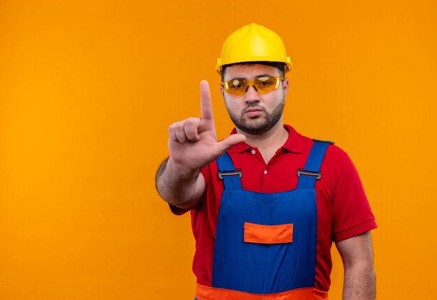 Uomo giovane costruttore in uniforme da costruzione e casco di sicurezza che mostra il dito indice con la faccia seria