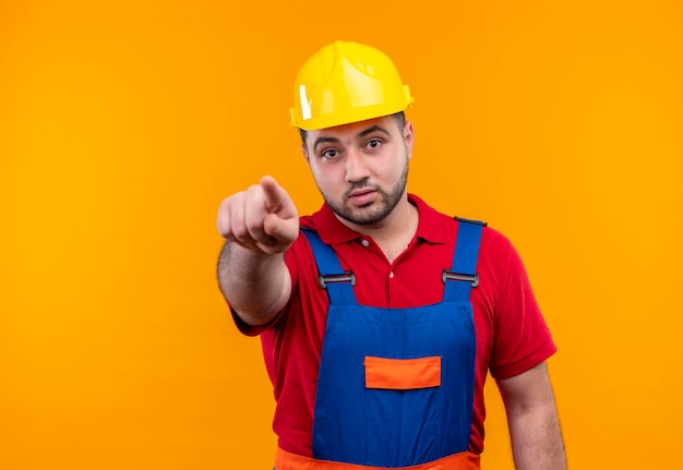 Uomo giovane costruttore in uniforme da costruzione e casco di sicurezza che punta con il dito indice alla telecamera che sembra sorpreso