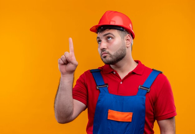 Uomo giovane costruttore in uniforme da costruzione e casco di sicurezza che punta con il dito indice in alto che sembra incerto