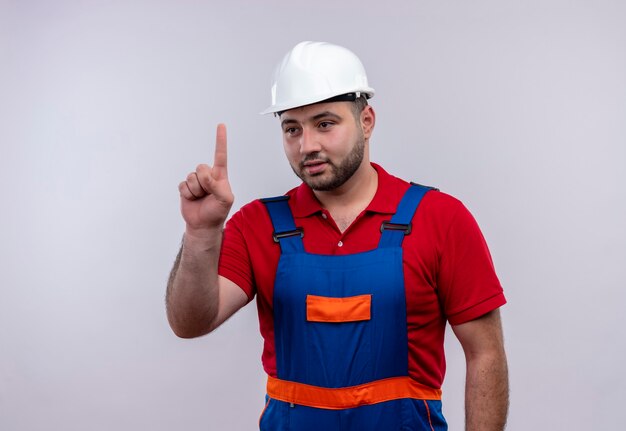 Uomo giovane costruttore in uniforme da costruzione e casco di sicurezza che punta con il dito indice in su guardando fiducioso