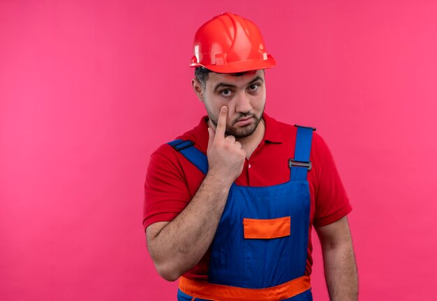 Uomo giovane costruttore in uniforme da costruzione e casco di sicurezza che punta con il dito indice per occhio guardandoti gesto