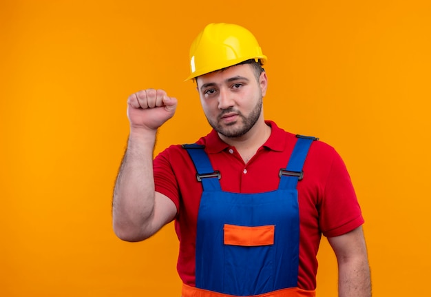 Uomo giovane costruttore in uniforme da costruzione e casco di sicurezza che stringe il pugno che guarda l'obbiettivo con la faccia seria