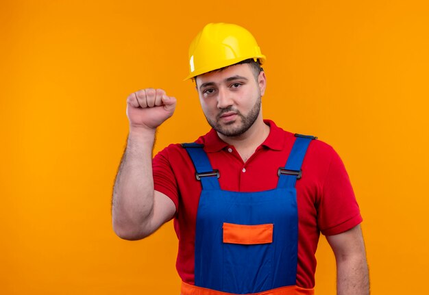 Uomo giovane costruttore in uniforme da costruzione e casco di sicurezza che stringe il pugno che guarda l'obbiettivo con la faccia seria