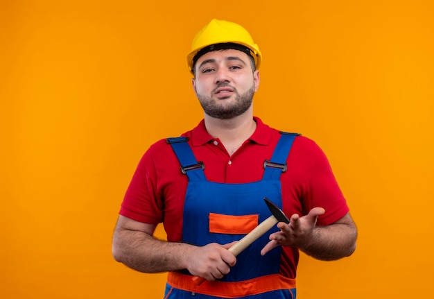 Uomo giovane costruttore in uniforme da costruzione e casco di sicurezza che tiene martello cercando fiducioso