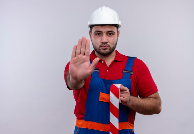 Uomo giovane costruttore in uniforme da costruzione e casco di sicurezza con la mano aperta che fa il fanale di arresto con la faccia seria