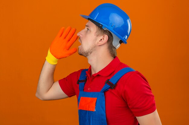 Uomo giovane costruttore in uniforme da costruzione e casco di sicurezza guardando in alto e gridando ad alta voce arrabbiato con le mani sopra la bocca in piedi lateralmente sul muro arancione isolato