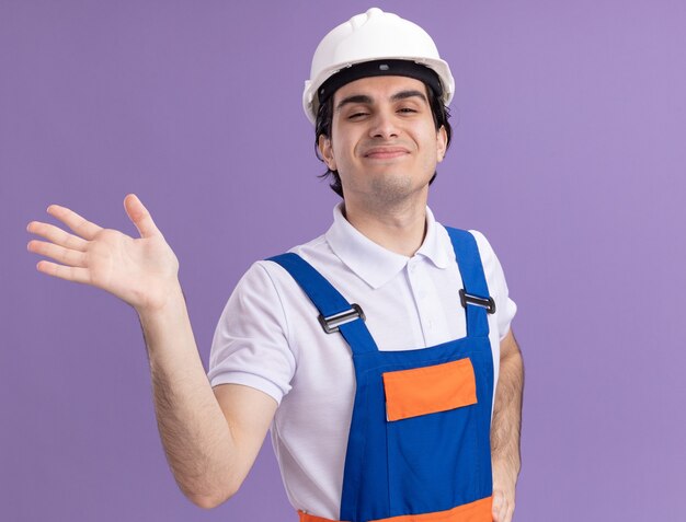 Uomo giovane costruttore in uniforme da costruzione e casco di sicurezza guardando la parte anteriore con la faccia felice in piedi sopra la parete viola