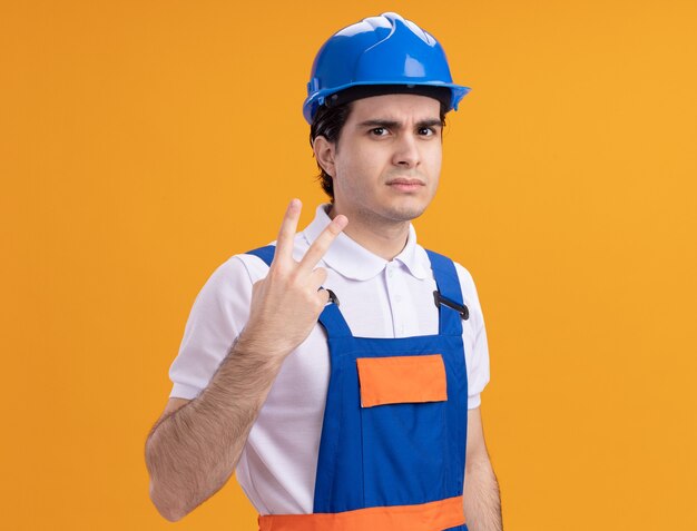 Uomo giovane costruttore in uniforme da costruzione e casco di sicurezza guardando la parte anteriore con la faccia seria che mostra il numero due in piedi sopra la parete arancione