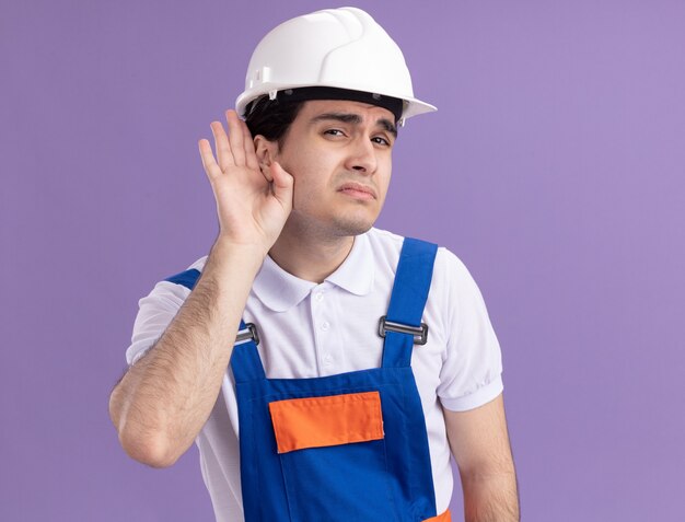 Uomo giovane costruttore in uniforme da costruzione e casco di sicurezza guardando la parte anteriore con la mano sull'orecchio cercando di ascoltare i pettegolezzi in piedi sopra il muro viola