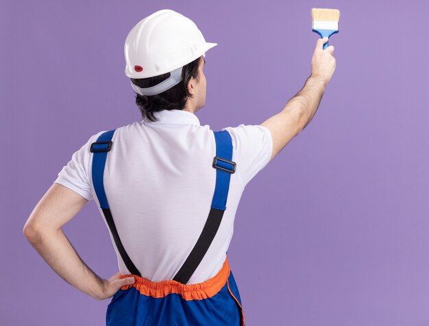 Uomo giovane costruttore in uniforme da costruzione e casco di sicurezza in piedi con il suo muro di vernice posteriore con rullo di vernice sul muro viola