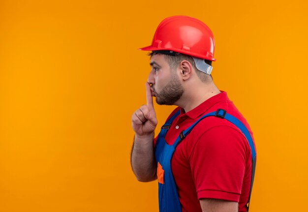 Uomo giovane costruttore in uniforme da costruzione e casco di sicurezza lateralmente facendo gesto di silenzio con il dito sulle labbra