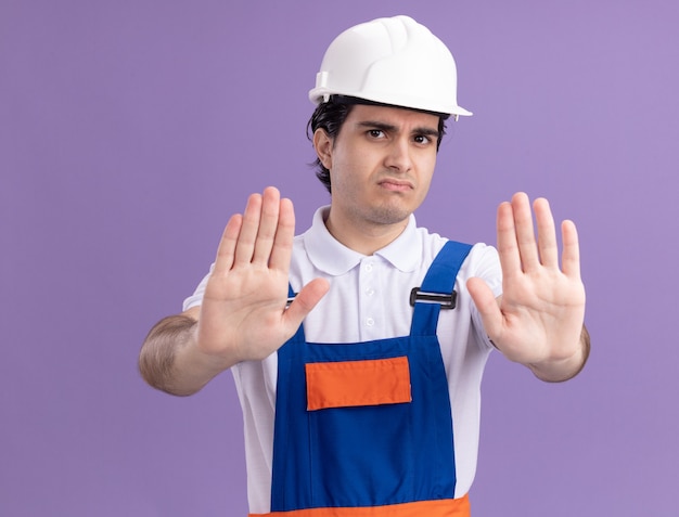 Uomo giovane costruttore in uniforme da costruzione e casco di sicurezza lookign davanti con faccia seria che fa gesto di arresto con le mani in piedi sopra la parete viola