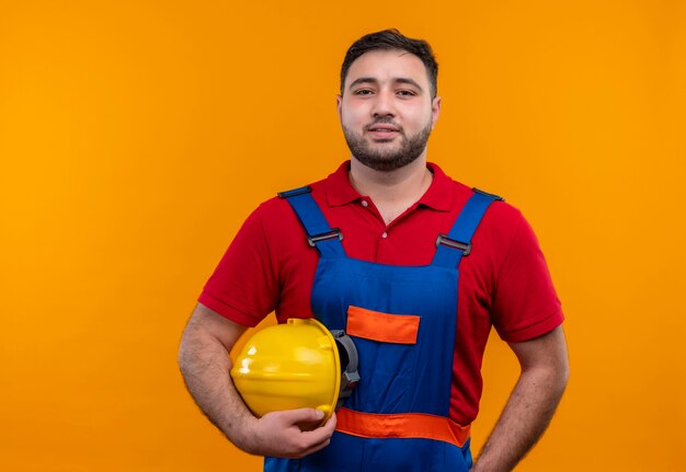 Uomo giovane costruttore in uniforme di costruzione che tiene il casco di sicurezza che guarda l'obbiettivo con un sorriso fiducioso