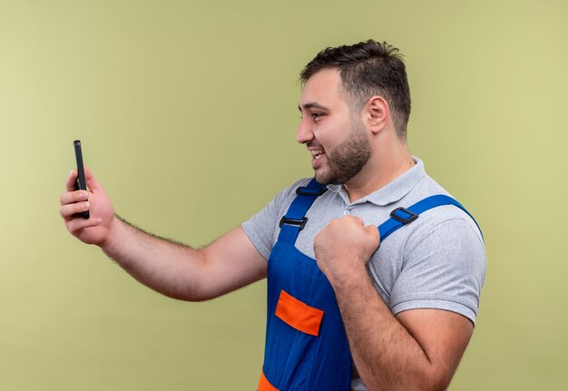 Uomo giovane costruttore in uniforme di costruzione che tiene smartphone pugno di serraggio felice ed uscito