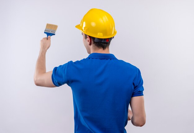 Uomo giovane costruttore indossa uniforme da costruzione e casco di sicurezza dipinge il muro con il pennello