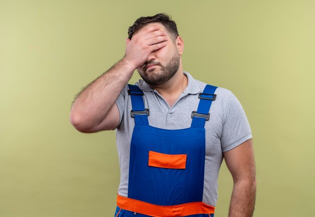 Uomo giovane costruttore infastidito in uniforme da costruzione che copre gli occhi con il braccio