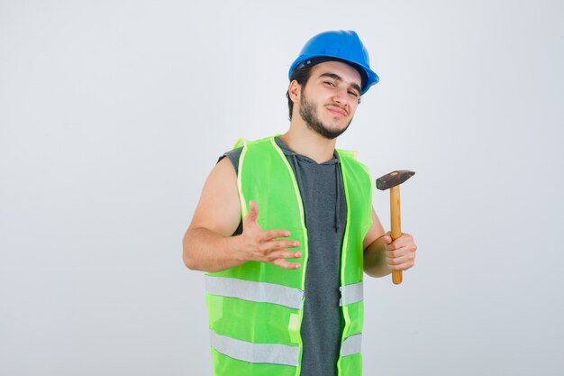 Uomo giovane costruttore tenendo un martello in uniforme e guardando fiducioso, vista frontale.