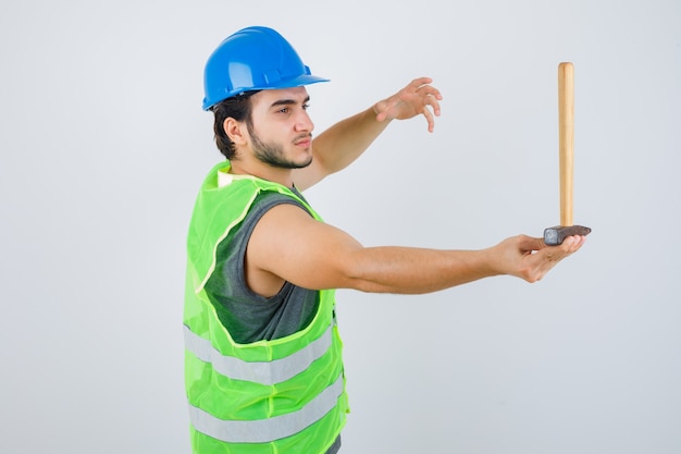 Uomo giovane costruttore tenendo un martello mentre si alza la mano in uniforme da lavoro e guardando attento, vista frontale.