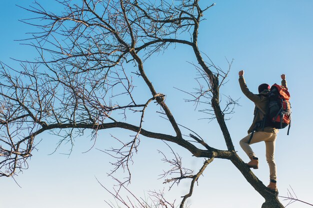 Uomo giovane hipster che viaggia con lo zaino, in piedi sull'albero contro il cielo, indossa una giacca calda, turista attivo, esplorando la natura nella stagione fredda