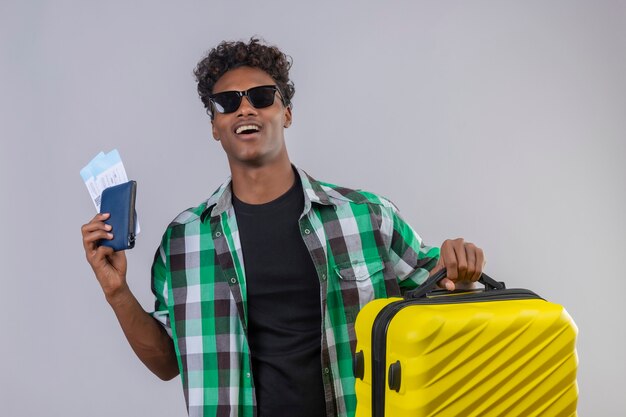 Uomo giovane viaggiatore afroamericano che indossa occhiali da sole neri in piedi con la valigia in possesso di biglietti aerei sorridendo allegramente positivo e felice su sfondo bianco