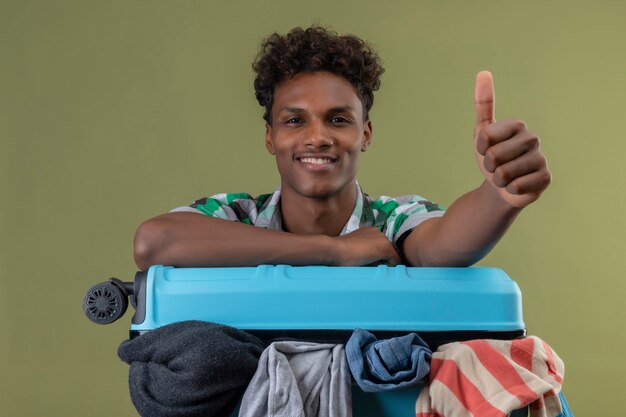 Uomo giovane viaggiatore afroamericano con la valigia piena di vestiti che guarda l'obbiettivo sorridente allegramente, positivo e felice, mostrando pollice in alto su sfondo verde