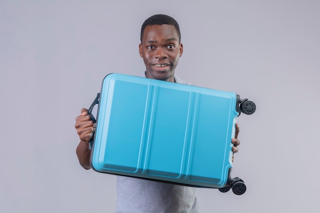 Uomo giovane viaggiatore afroamericano in camicia di polo grigia che tiene la valigia blu che sembra sorpreso e felice