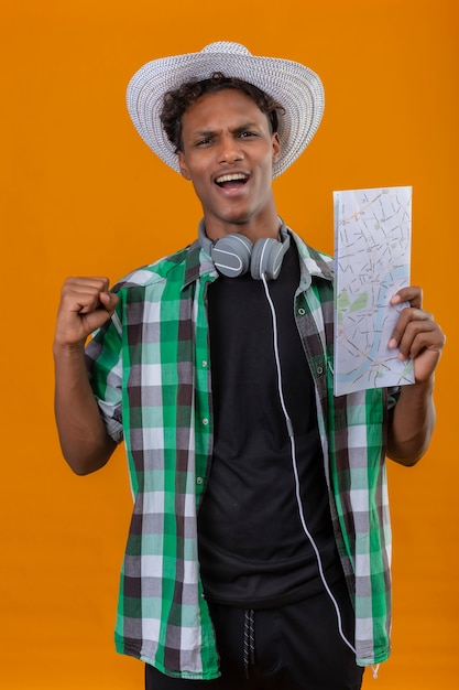 Uomo giovane viaggiatore afroamericano in cappello estivo con le cuffie che tengono mappa alzando il pugno uscito e felice alzando il pugno gioendo del suo successo in piedi su sfondo arancione