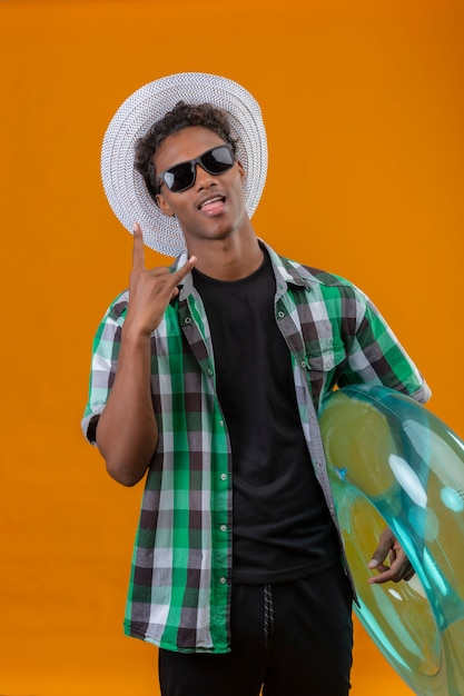 Uomo giovane viaggiatore afroamericano in cappello estivo indossando occhiali da sole neri che tiene anello gonfiabile facendo segno di roccia con la lingua fuori, divertendosi, guardando la telecamera sopra la parte posteriore arancione