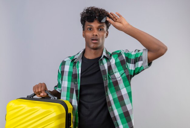 Uomo giovane viaggiatore afroamericano stupito che tiene la valigia che toglie gli occhiali da sole dalla condizione di stupore