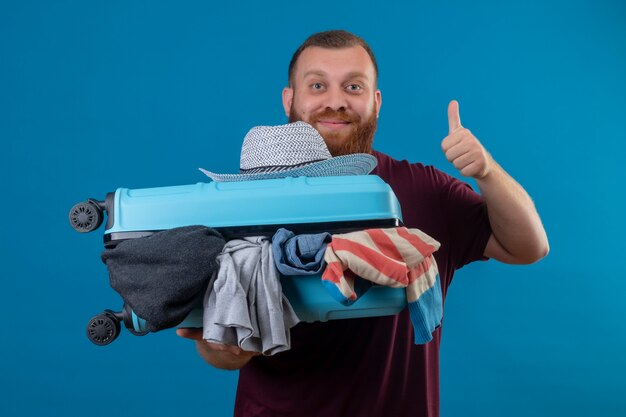 Uomo giovane viaggiatore barbuto che tiene la valigia piena di vestiti sorridente ottimista e felice che mostra i pollici in su