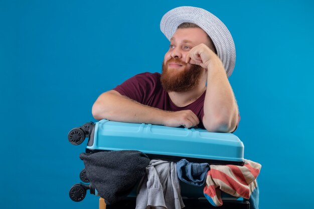 Uomo giovane viaggiatore barbuto in cappello estivo con la valigia piena di vestiti sorridente ottimista e felice che osserva da parte