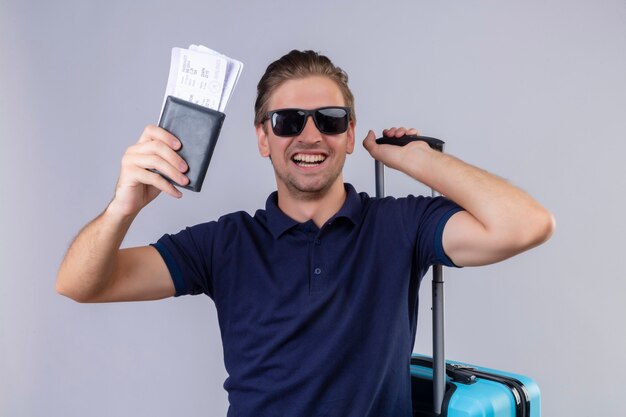 Uomo giovane viaggiatore bello che indossa occhiali da sole neri in possesso di biglietti aerei in piedi con la valigia felice ed è uscito sorridendo allegramente su sfondo bianco