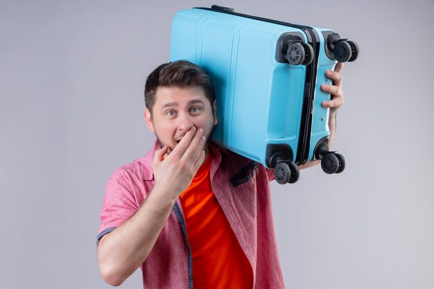 Uomo giovane viaggiatore bello che tiene la valigia blu sorpreso e stupito che copre la bocca con la mano in piedi sul muro bianco