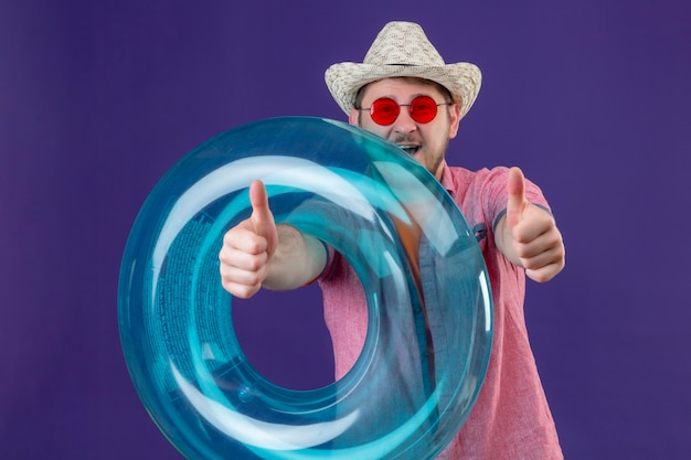 Uomo giovane viaggiatore bello in cappello estivo con anello gonfiabile che mostra i pollici in su