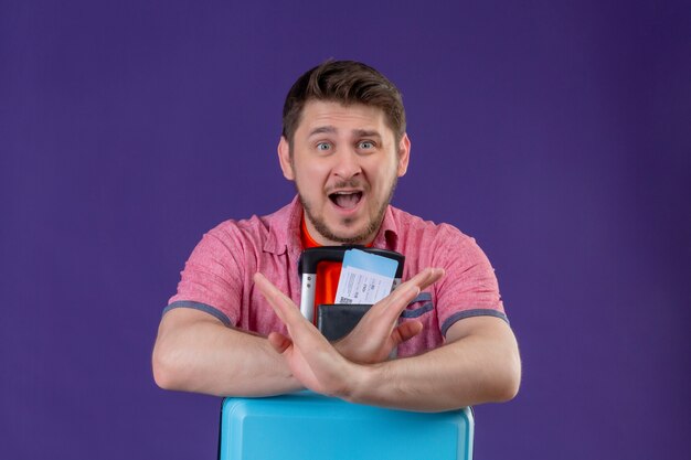 Uomo giovane viaggiatore che tiene la valigia e biglietti aerei guardando la telecamera stupito facendo gesto di arresto attraversando le mani in piedi su sfondo viola